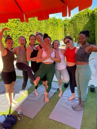 Eight women in activewear smiling and balancing on yoga mats in an outdoor poolside yoga class under a bright orange umbrella on artificial turf with a tall green hedge backdrop