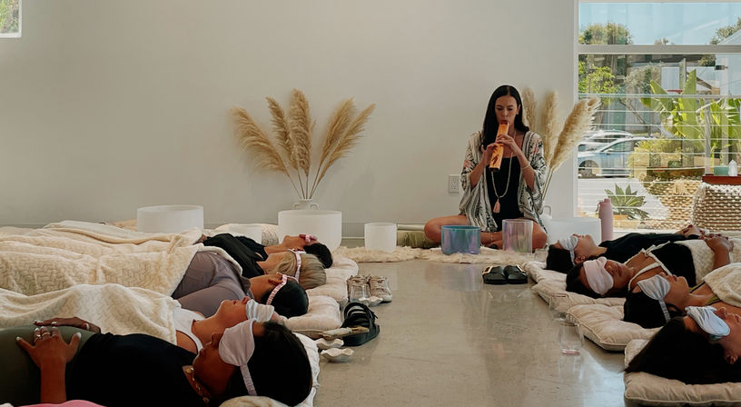Group sound bath in a bright wellness studio: participants recline on mats with eye masks and cozy blankets while a facilitator plays a wooden flute beside crystal singing bowls, pampas grass and large windows in the background.