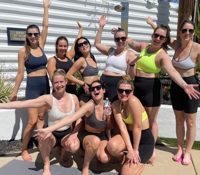Sunny poolside group of smiling women in athletic wear and sunglasses posing on a yoga mat with palm trees and a white privacy fence in the background.