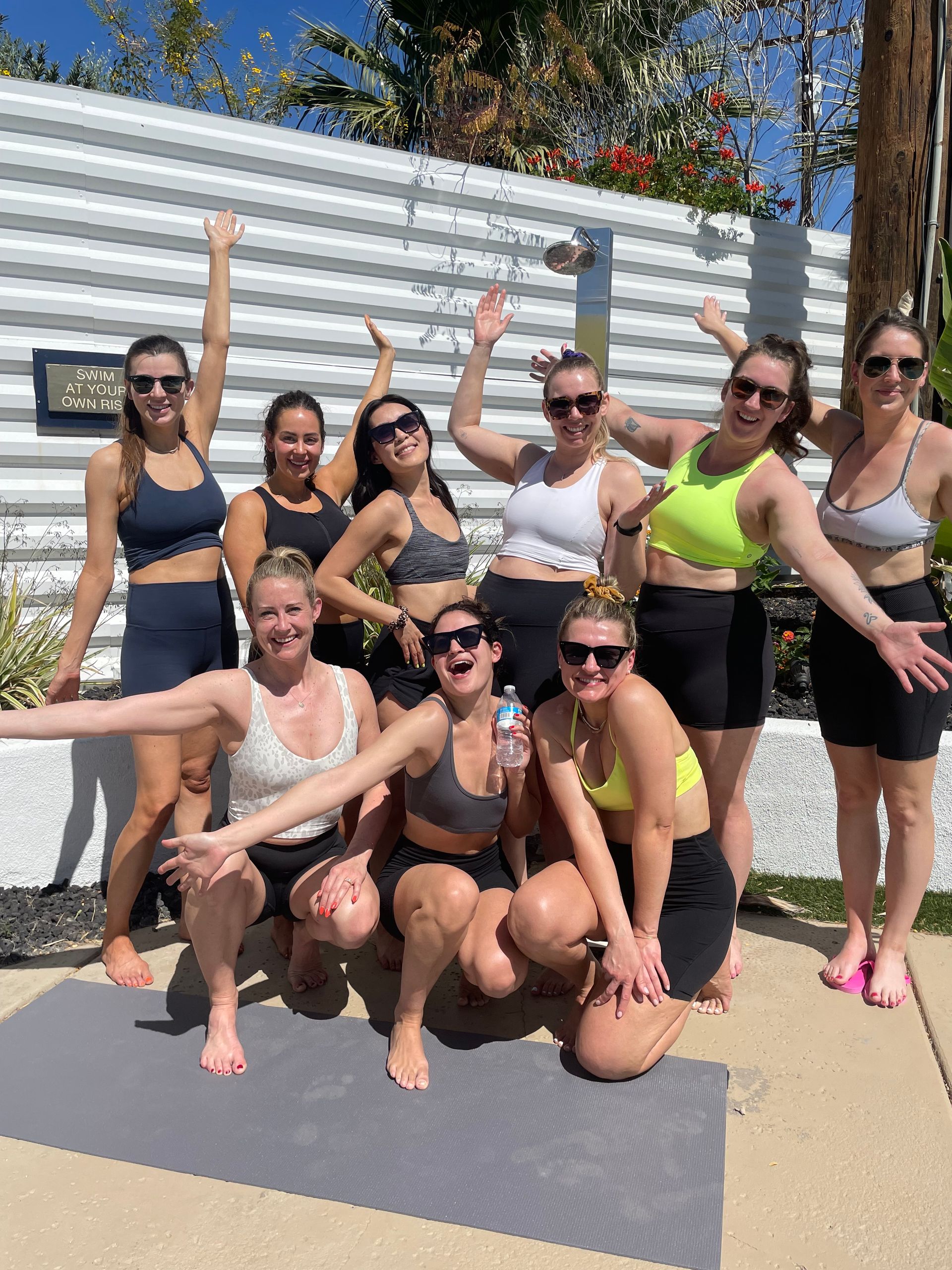 Sunny poolside group of smiling women in athletic wear and sunglasses posing on a yoga mat with palm trees and a white privacy fence in the background.