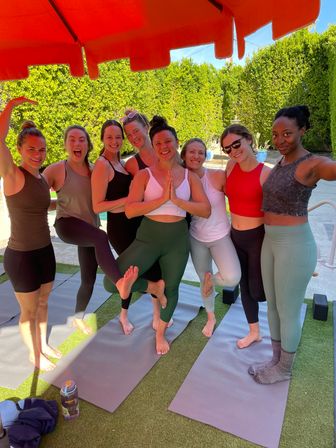Eight women practicing yoga on mats under a red umbrella on a sunlit backyard patio, smiling and striking tree poses.