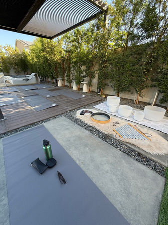 Shaded backyard patio set for outdoor yoga and sound‑healing — gray yoga mats on wood and concrete, crystal singing bowls and chimes on a faux‑fur rug, lounge chair, water bottle and bamboo privacy hedge.