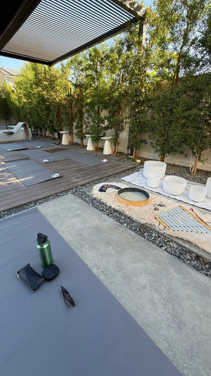 Shaded backyard patio set for outdoor yoga and sound‑healing — gray yoga mats on wood and concrete, crystal singing bowls and chimes on a faux‑fur rug, lounge chair, water bottle and bamboo privacy hedge.