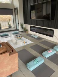 Sunlit modern meditation and yoga room with gray mats and teal bolsters arranged around crystal singing bowls on a white blanket, wall TV above a linear electric fireplace and large windows with potted cacti.