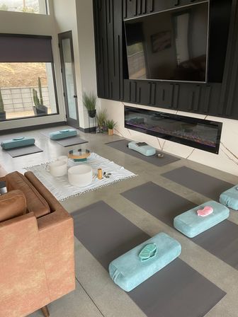 Sunlit modern meditation and yoga room with gray mats and teal bolsters arranged around crystal singing bowls on a white blanket, wall TV above a linear electric fireplace and large windows with potted cacti.