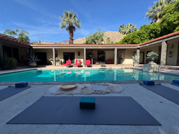 Sunlit desert courtyard with turquoise swimming pool surrounded by a single-story colonnade, red lounge chairs, palm trees and rocky hills; yoga mats and blocks set up on the pool deck for an outdoor session.