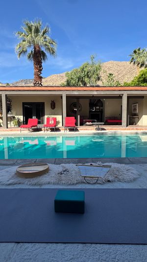 Sunlit desert courtyard with turquoise swimming pool surrounded by a single-story colonnade, red lounge chairs, palm trees and rocky hills; yoga mats and blocks set up on the pool deck for an outdoor session.