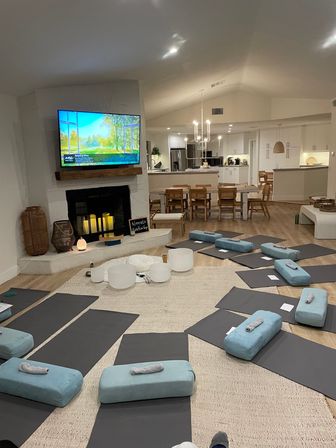 Open-plan living room staged for a group yoga class with black mats, teal bolster pillows and rolled towels on a large rug, crystal singing bowls by a fireplace, TV above, and a bright white kitchen and dining area in the back.