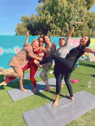 Five women in activewear laughing and posing barefoot on yoga mats during a sunny outdoor yoga session in front of a colorful mural and trees.