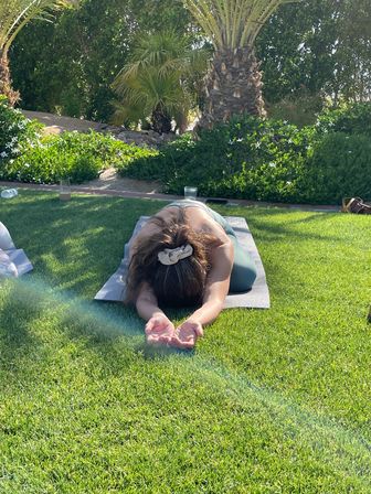 Person in a child's pose on a yoga mat on a sunlit green lawn, surrounded by palm trees and flowering shrubs in a tropical garden setting