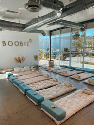 Sunlit wellness studio set for a group sound-bath meditation with rows of cushioned floor mats, teal bolsters, rolled towels, crystal singing bowls and pampas grass beside floor-to-ceiling windows overlooking sunlit hills and a parking area.