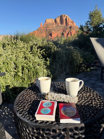 Two steaming coffee mugs, a sage smudge stick and oracle card deck on a round patio table with lounge chair, desert shrubs and a red‑rock butte under a clear blue sky in Sedona.
