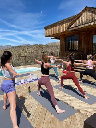 Sun-drenched outdoor yoga class on a wooden deck beside a plunge pool at a desert mountain retreat, participants in colorful activewear holding Warrior II pose against a bright blue sky.