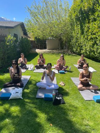 Group of women in a backyard yoga class sitting on mats on a sunny green lawn, smiling and enjoying bowls after practice beneath leafy trees.