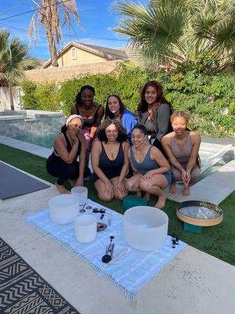 Seven women smiling poolside in a sunny backyard with palm trees, gathered around crystal singing bowls, a drum and wellness tools on a towel for an outdoor sound‑bath meditation session.