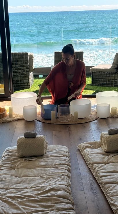 Sunlit oceanfront sound bath — a person playing crystal singing bowls inside a beachfront room with candles and floor cushions, waves visible through glass doors