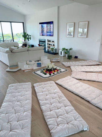 Sunlit modern living room set for group meditation with white floor mats around a floral centerpiece, white sectional sofa, indoor plants, TV above fireplace and balcony view