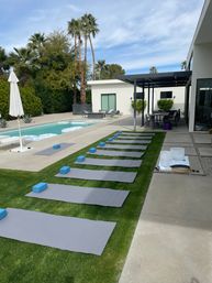 Poolside yoga setup in a modern desert-style backyard: gray mats and blue blocks lined on the lawn, white sound bowls, swimming pool, palm trees and covered patio.