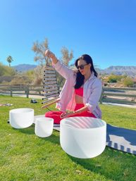 Woman in pink activewear and sunglasses seated on a yoga mat holding a metal chime above white crystal singing bowls on a green lawn — outdoor sound healing in a desert park with palm trees, mountains, and clear blue sky