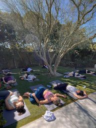 Outdoor yoga class in a sunny backyard with participants on mats resting in reclined poses beneath a large tree on green lawn.
