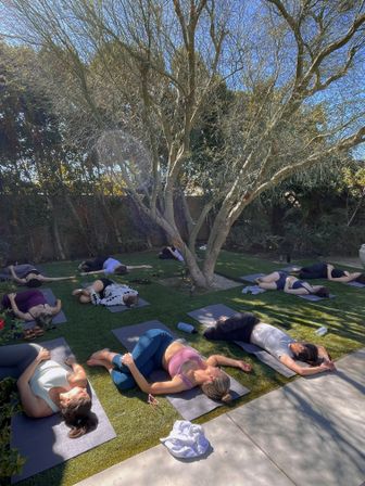Outdoor yoga class in a sunny backyard with participants on mats resting in reclined poses beneath a large tree on green lawn.