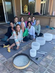 Group of women seated on yoga mats on a sunlit backyard patio for a sound bath session, with white crystal singing bowls, mallets and a frame drum arranged on the mats.