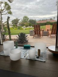 Crystal singing bowls on a mat by a floor-to-ceiling window, overlooking a palm-lined backyard with agave and cactus, green lawn, rock landscaping and wooden patio chairs under a cloudy sky.