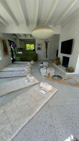 Bright modern living room set up for a sound bath/meditation: beige floor mats with rolled blankets and eye masks, crystal singing bowls and instruments on a fur runner, white brick fireplace, large paper lantern pendant and a green sofa near windows.