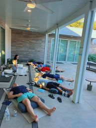 Group reclining meditation on a modern covered patio — participants on mats with bolsters, water bottles and notebooks while an instructor sits near crystal singing bowls by sliding glass doors.