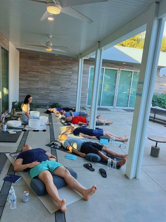 Group reclining meditation on a modern covered patio — participants on mats with bolsters, water bottles and notebooks while an instructor sits near crystal singing bowls by sliding glass doors.
