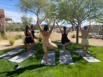 Five women in activewear doing tree pose on yoga mats in a sunny desert garden by a pool, shaded by trees and surrounded by agave plants and rocky hills.