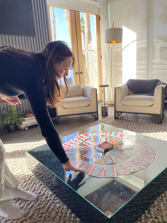 Person leaning over a glass coffee table arranging red-backed playing cards in a spiral on a chunky rug in a sunlit modern living room with beige armchairs and tall wooden glass doors