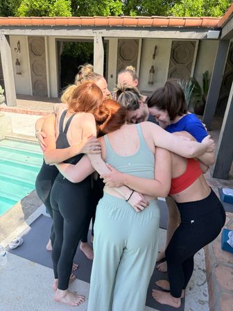 Seven women in activewear sharing a warm group hug on yoga mats beside a sunny backyard pool, outdoor wellness retreat vibe