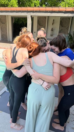 Seven women in activewear sharing a warm group hug on yoga mats beside a sunny backyard pool, outdoor wellness retreat vibe