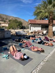 Outdoor yoga retreat: participants in savasana lying on mats with blocks and straps on a sunny desert patio, palm tree and terracotta-roof cottage in the foreground and rocky mountains under a blue sky in the background.