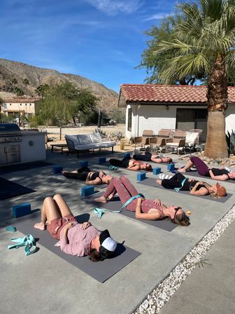 Outdoor yoga retreat: participants in savasana lying on mats with blocks and straps on a sunny desert patio, palm tree and terracotta-roof cottage in the foreground and rocky mountains under a blue sky in the background.
