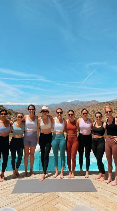 Ten women in colorful activewear standing barefoot on a wooden pool deck with a yoga mat and pool, smiling under a clear blue sky with snow-capped desert mountains in the background — sunny outdoor yoga/fitness retreat scene.