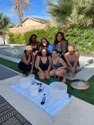 Seven women gathered in a sunny suburban backyard by a pool and palm trees, smiling and posing around crystal singing bowls and sound‑healing tools arranged on a striped blanket.