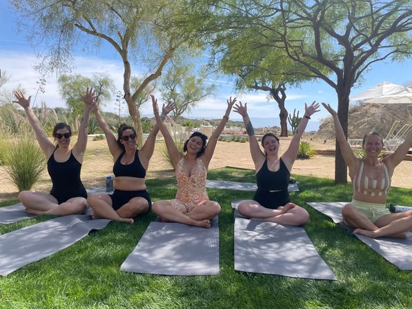 Five women seated cross-legged on yoga mats with arms raised, smiling during outdoor yoga on a green lawn under trees by a sunny desert poolside landscape.
