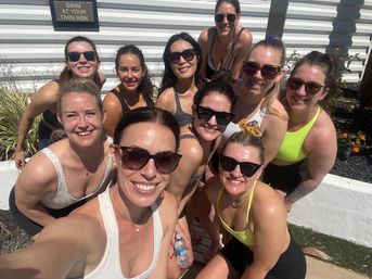 Group selfie of eleven women in athletic swimwear and sunglasses smiling on a sunny outdoor pool deck beside a "Swim at your own risk" sign