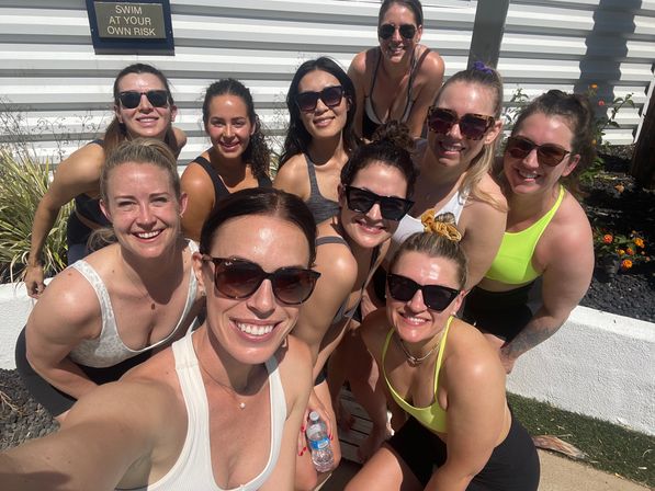 Group selfie of eleven women in athletic swimwear and sunglasses smiling on a sunny outdoor pool deck beside a "Swim at your own risk" sign