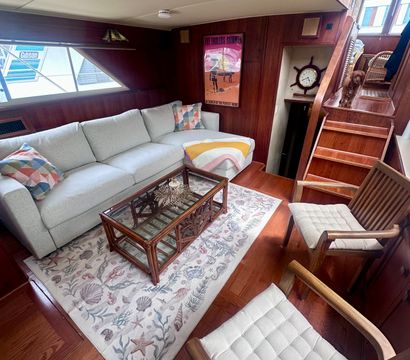 Cozy yacht salon — light gray sectional with colorful pillows, rattan glass coffee table on a seashell rug, wood-paneled walls, nautical clock and a small dog on the stairs.