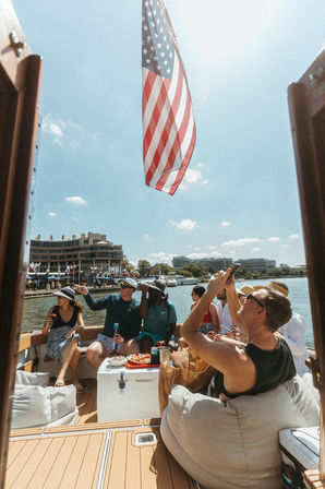 Summer boat party with friends lounging under a large American flag at a sunny waterfront marina, yachts and buildings along the harbor.