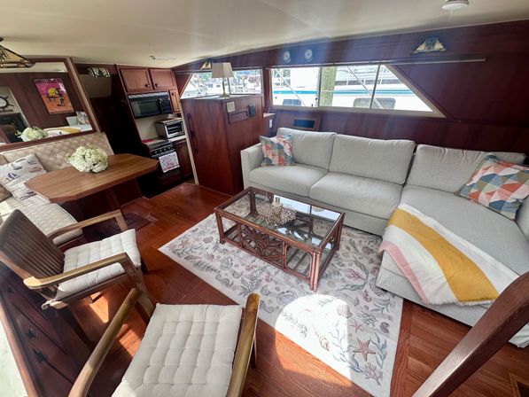 Bright yacht salon interior with light-gray L-shaped sofa, colorful throw pillows and striped blanket, glass-top rattan coffee table on a seashell-pattern rug, wooden dining nook and compact galley with stove and microwave, wood-paneled walls and marina view through long windows.