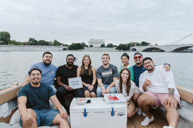Cheerful group of friends on a boat cruise along the Washington, D.C. waterfront with the Lincoln Memorial and bridge in the background, one holding a small 'Welcome Aboard' sign.