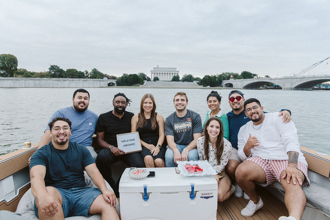 Cheerful group of friends on a boat cruise along the Washington, D.C. waterfront with the Lincoln Memorial and bridge in the background, one holding a small 'Welcome Aboard' sign.