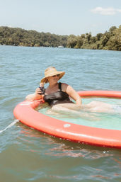 Smiling woman in a black swimsuit and wide-brim sun hat lounging on a red inflatable ring with sunglasses and a canned drink, floating on a sunny lake with tree-lined shore — relaxed summer boating scene
