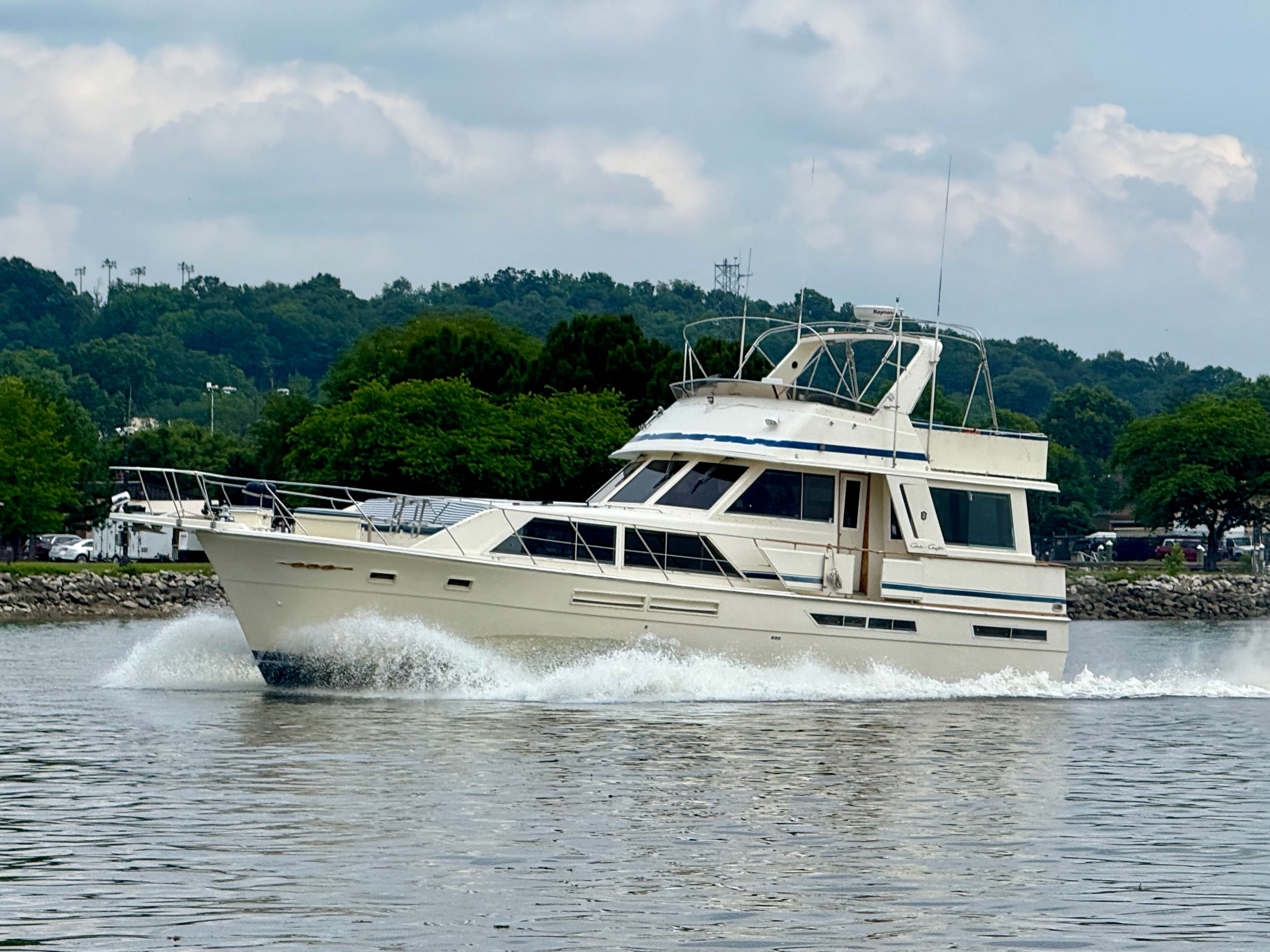 Cream-colored motor yacht cutting through calm harbor water, bow wake spraying past a tree-lined shoreline under a cloudy sky.