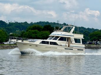 Cream-colored motor yacht cutting through calm harbor water, bow wake spraying past a tree-lined shoreline under a cloudy sky.