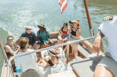 Group of friends laughing and sipping drinks on a sunny summer boat cruise, seated on a small motorboat over calm green water with an American flag at the stern.